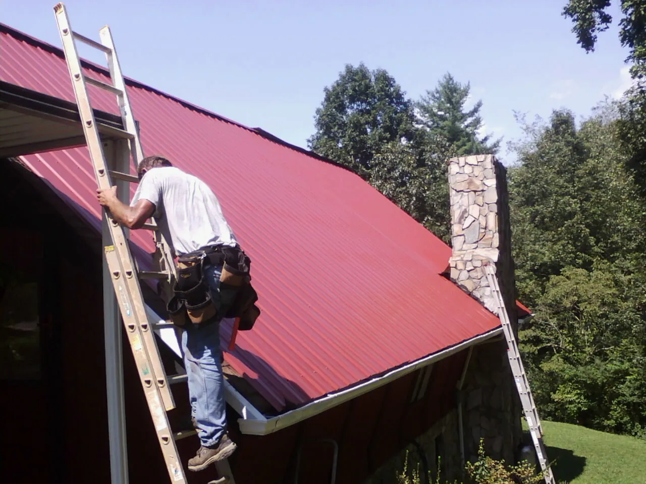 Roofer on a ladder beside a red metal roof and stone chimney — Soffit & Fascia Repair by Roofing Brothers in Etowah